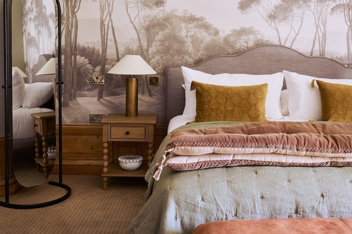 A bedroom with a bed, bedside table, lamp, and a wall with a nature-themed mural.