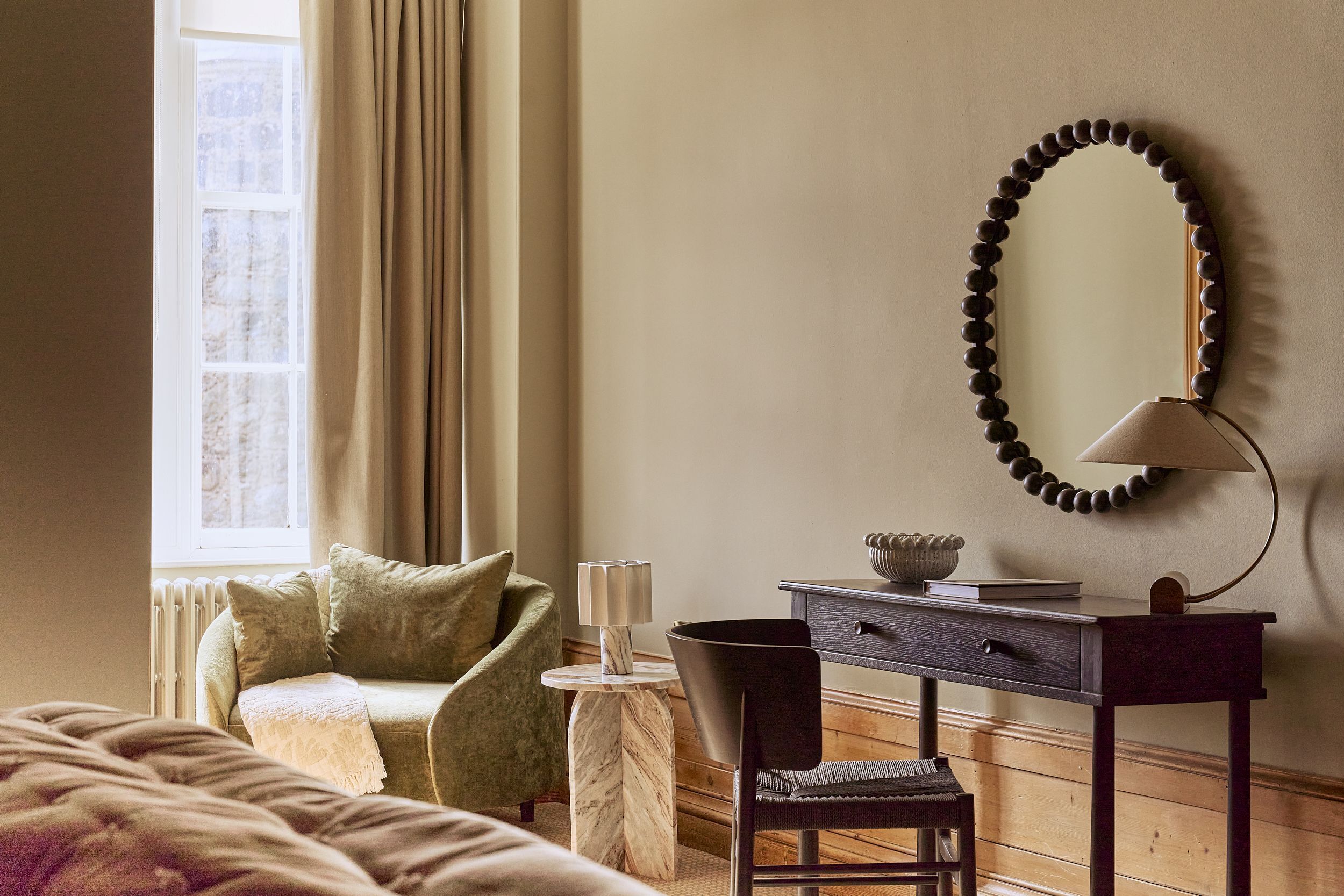 A room with a bed, a desk with a lamp, a chair, a mirror, and a window with curtains.