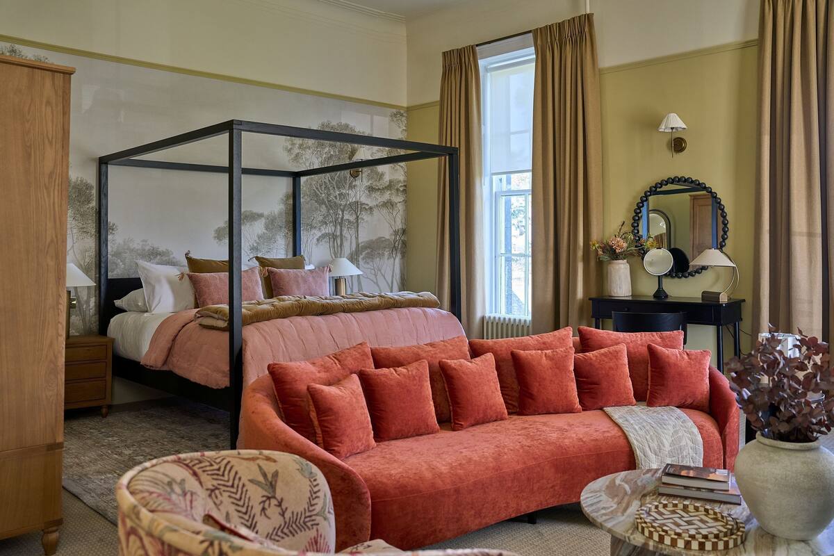 A bedroom with a four-poster bed, a red sofa, a wooden nightstand, and a mirror.