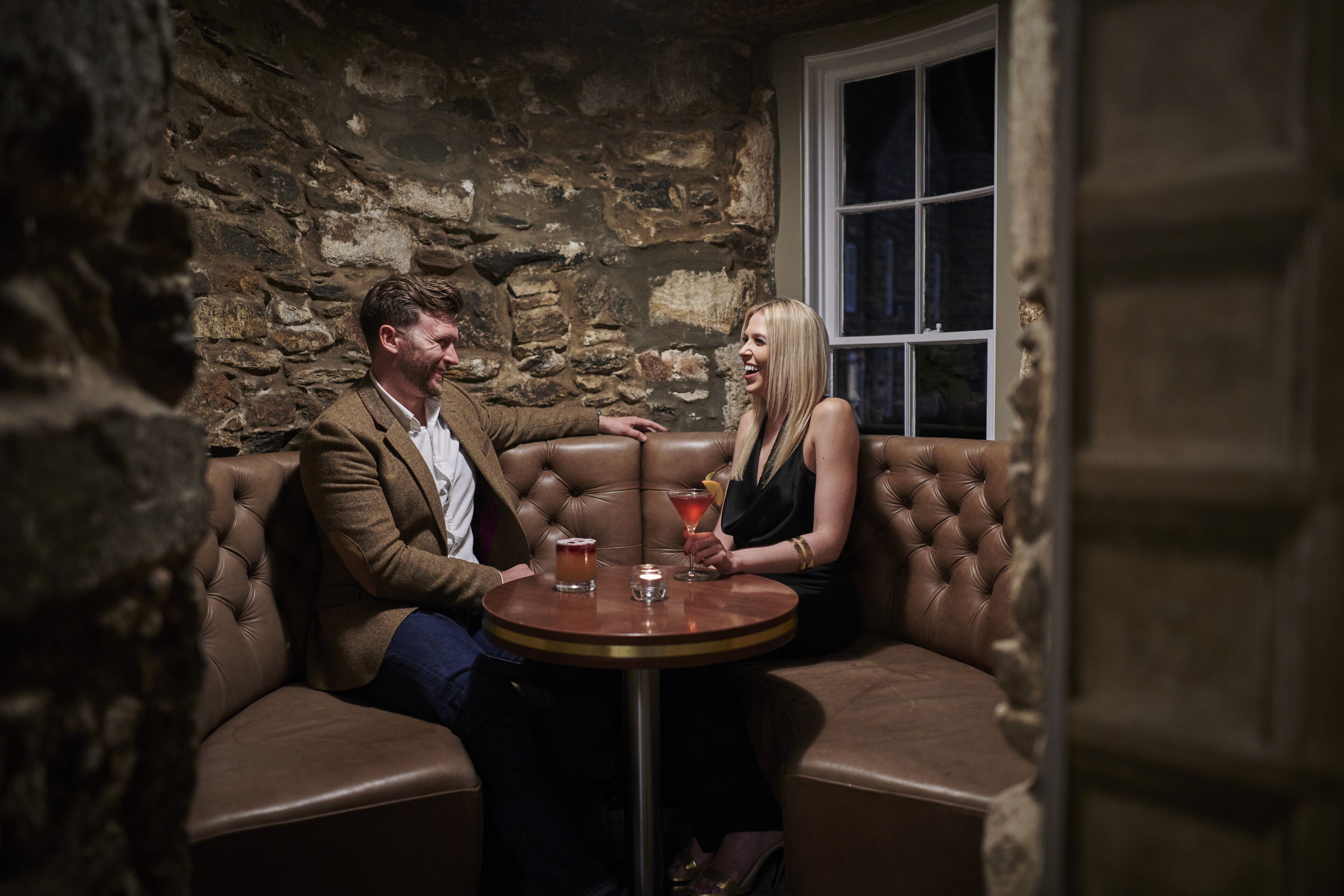 A couple seated on a brown leather sofa in a stone-walled room with a window.