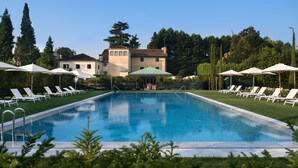 Una piscina rectangular con superficie de agua azul cristalina, rodeada de sillas de descanso y sombrillas blancas, con un edificio y árboles al fondo.