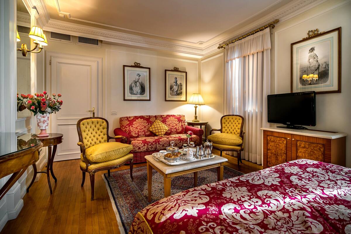 Habitación de hotel con colcha de diseño rojo y blanco, sofá con estampado floral rojo y dorado, dos sillones amarillos, una mesa de centro de madera con una bandeja de alimentos para el desayuno, un televisor sobre un mueble de madera y cuadros en las paredes.
