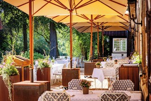 Een terras met tafels en stoelen, omringd door bomen en overdekt met grote parasols.