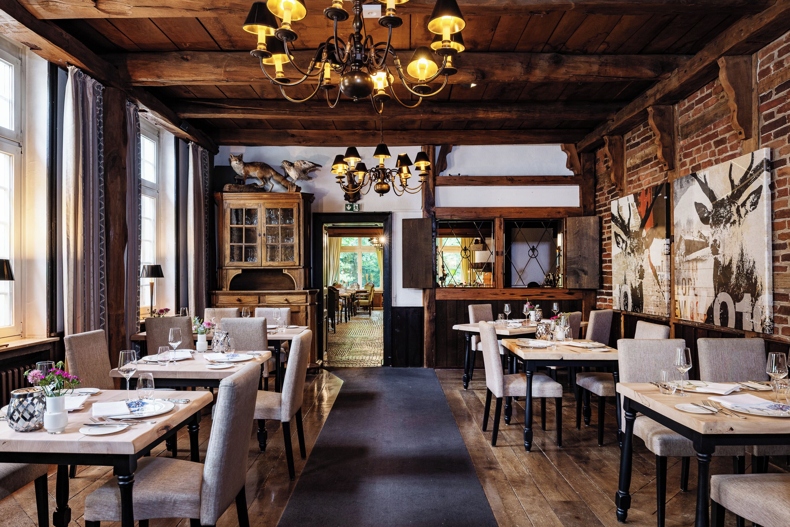 A restaurant interior with wooden beams, chandeliers, and a brick wall with a deer painting. Tables are set for dining.