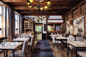 L’intérieur d’un restaurant présente des poutres en bois, des lustres et un mur de briques orné d’un tableau représentant un cerf. Les tables sont dressées pour le service.