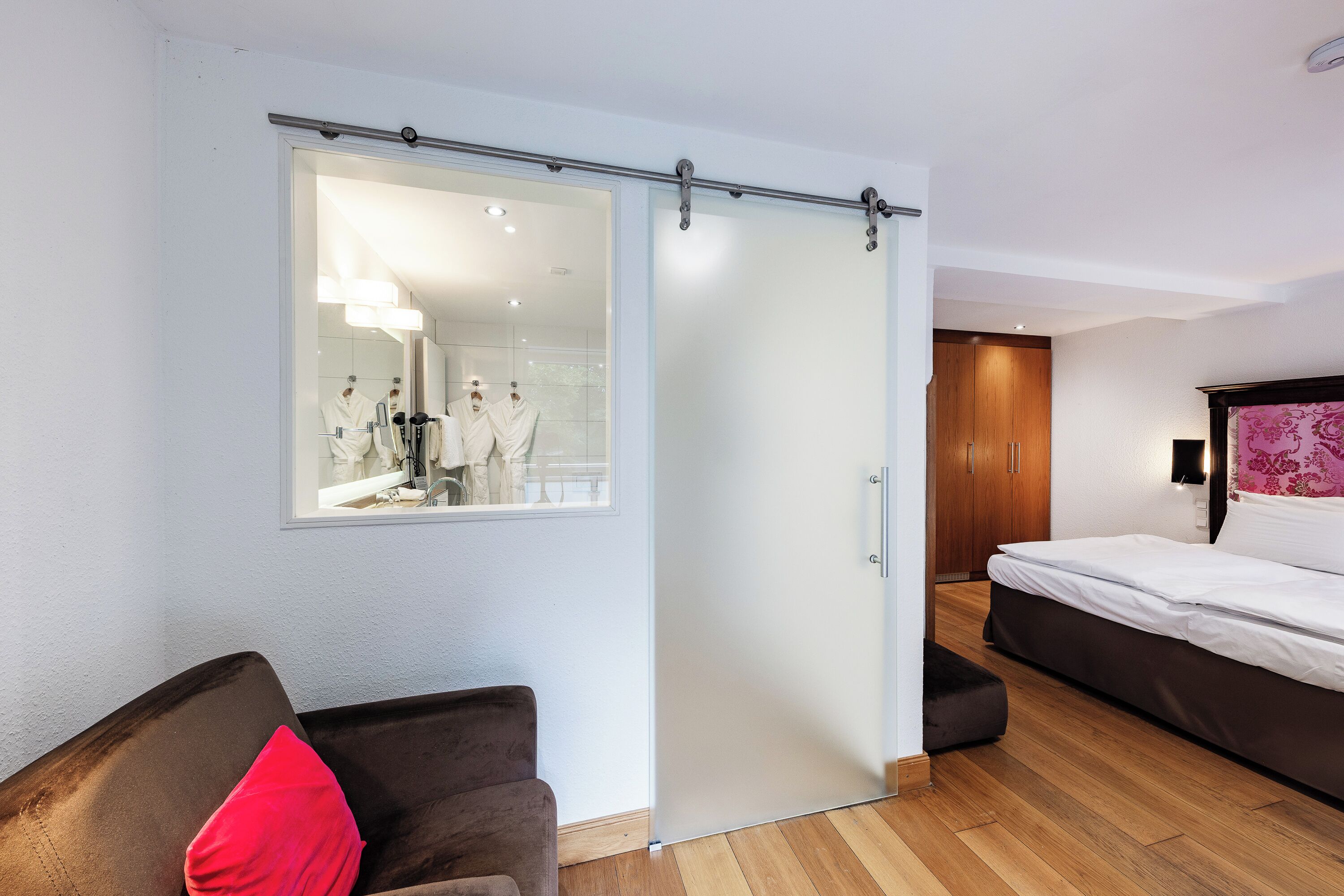 Hotelkamer met een schuifdeur naar een badkamer, een bed, een stoel met een rode kussen en een houten vloer.