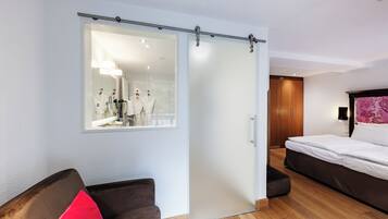 Hotelkamer met een schuifdeur naar een badkamer, een bed, een stoel met een rode kussen en een houten vloer.