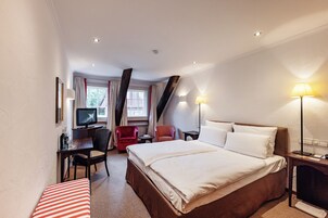 A hotel room with a bed, a desk with a computer, a chair, a television, and a lamp.