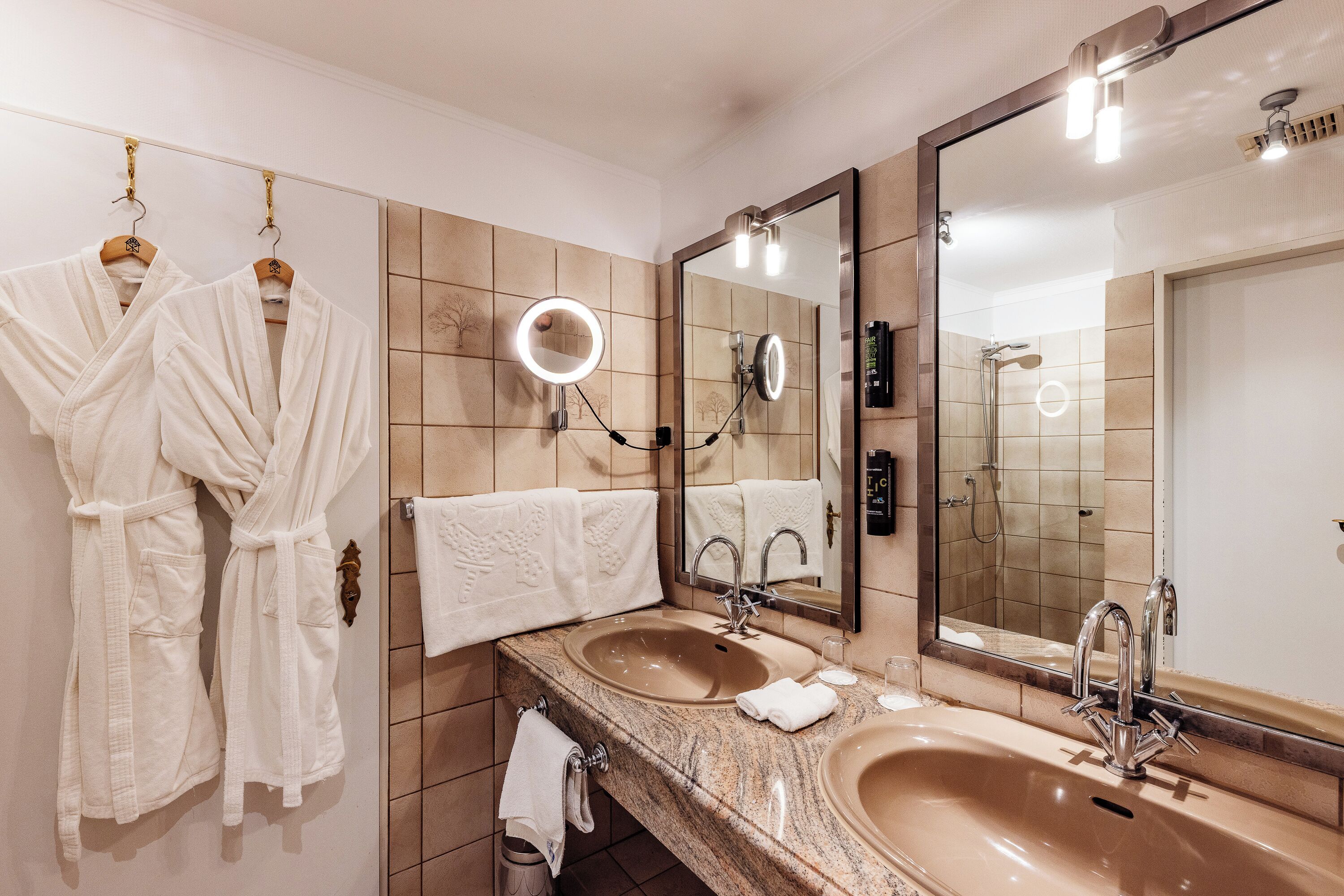 Un baño con dos lavamanos, un espejo grande y un perchero con batas.
