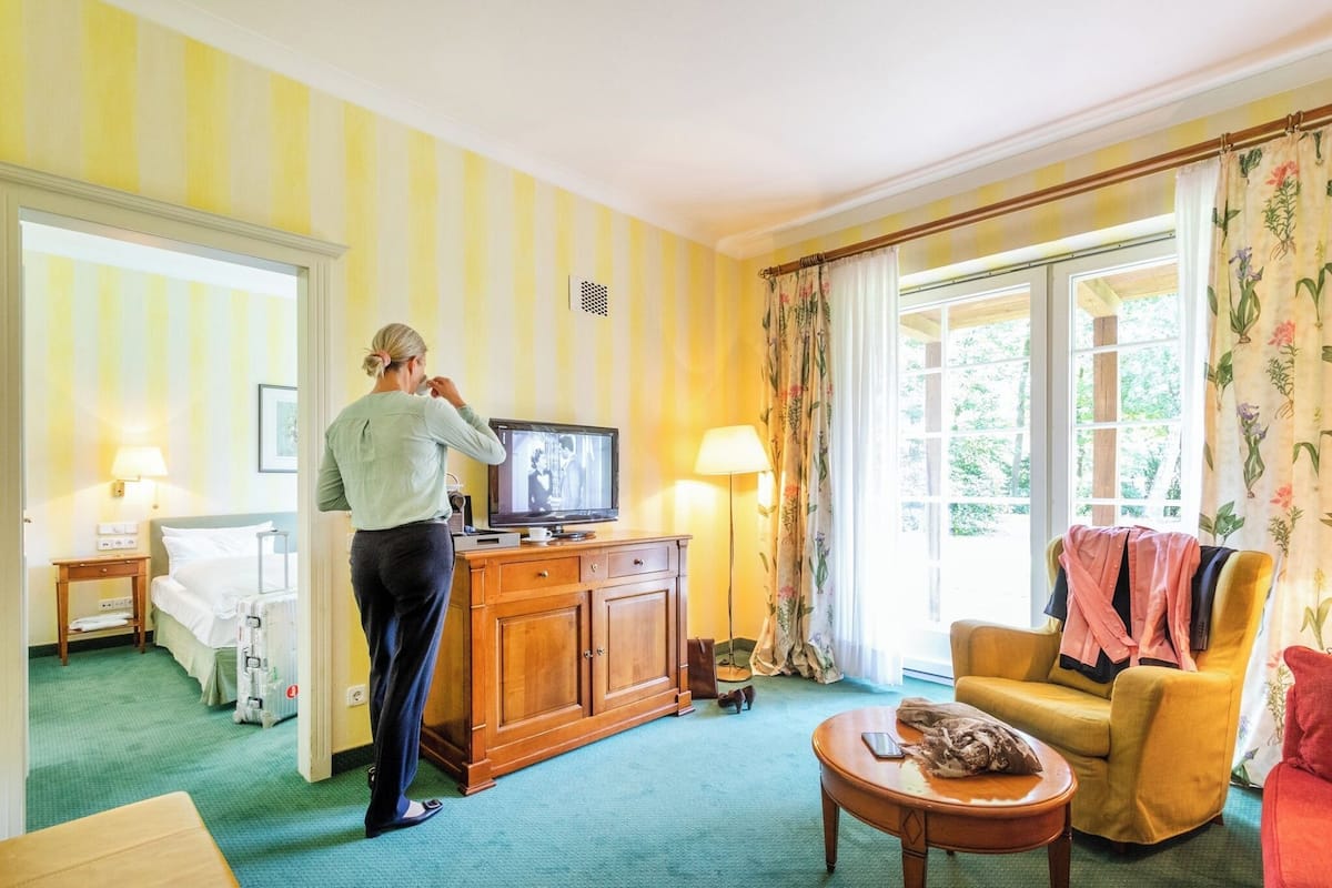 Une chambre d’hôtel avec un lit, une télévision, une armoire en bois, un fauteuil jaune et une fenêtre avec des rideaux.