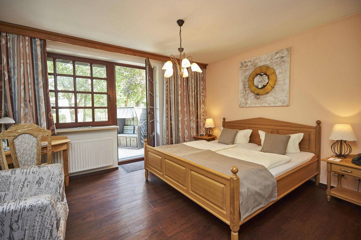 A bedroom with a wooden bed, a nightstand with a lamp, a chair, a window with curtains, and a wall with a decorative plate.