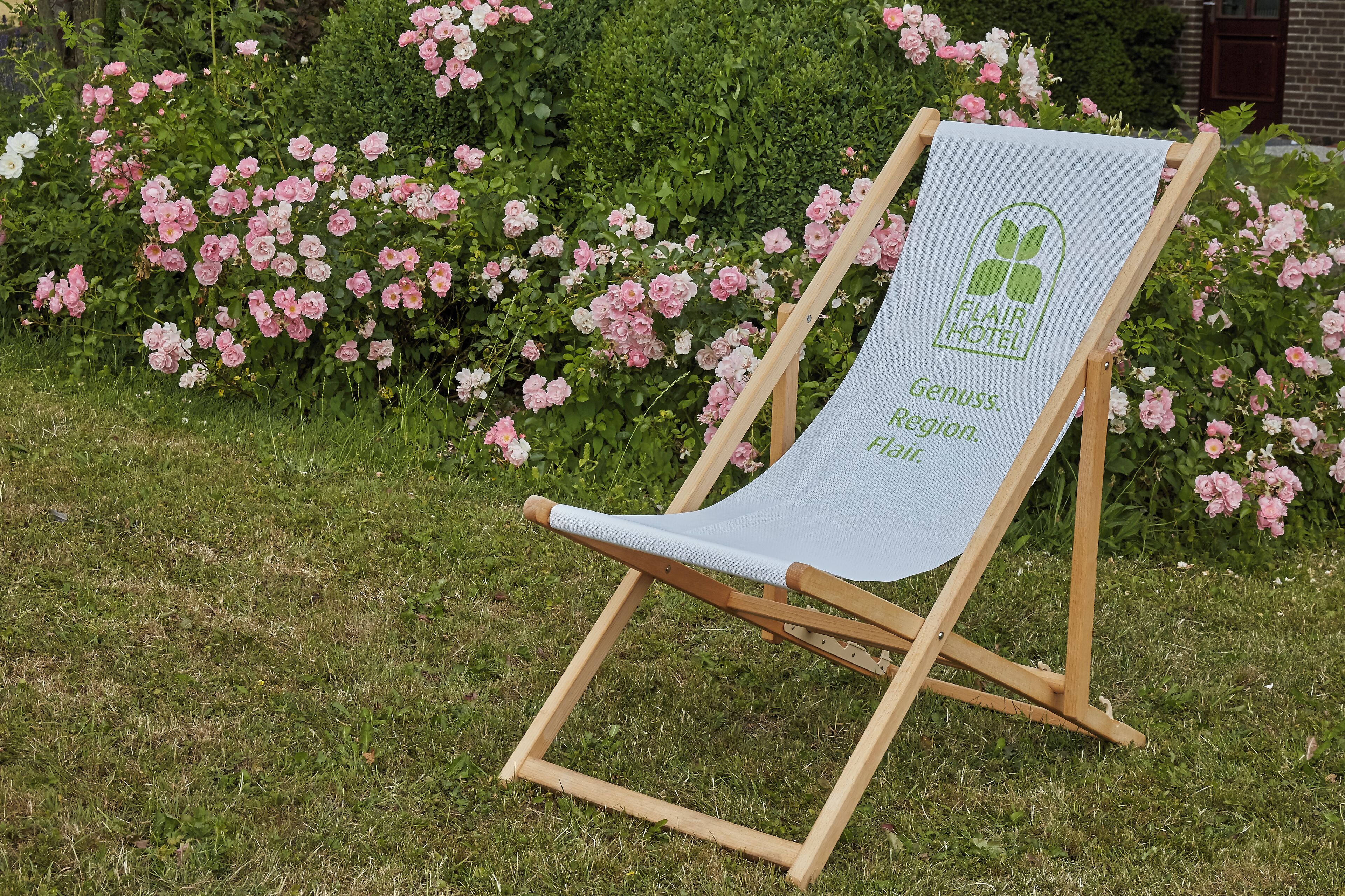 A wooden lounge chair with a white cushion and a green logo, placed outdoors with pink flowers in the background.