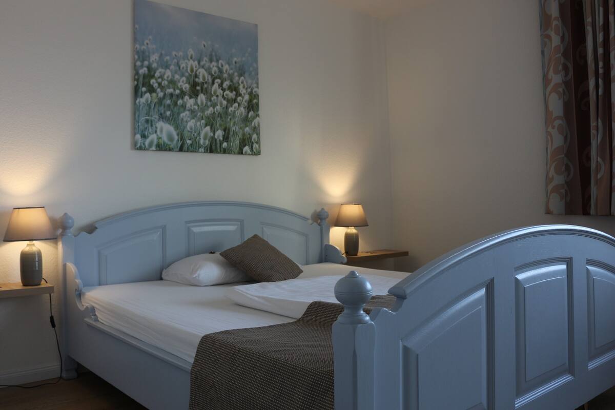 A bedroom with a bed, two bedside tables with lamps, a wall-mounted painting, and a window with curtains.