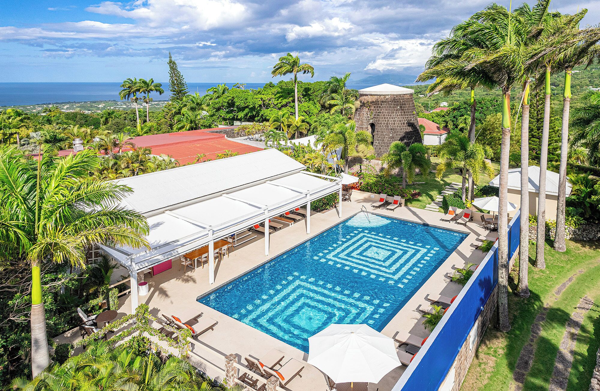 A swimming pool with a geometric tile pattern, surrounded by lounge chairs and umbrellas, set against a backdrop of lush greenery and a distant view of the ocean.