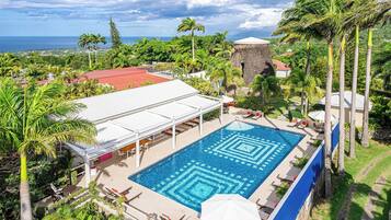 A swimming pool with a geometric tile pattern, surrounded by lounge chairs and umbrellas, set against a backdrop of lush greenery and a distant view of the ocean.