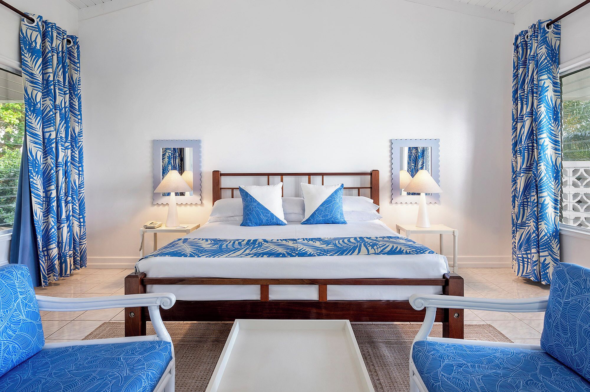A bedroom with a wooden bed, two bedside tables with lamps, a white nightstand, and a view of greenery outside.