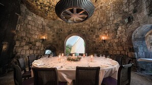 Una mesa redonda para seis personas, preparada con velas, rodeada de sillas de mimbre, en una habitación de paredes de piedra con un gran ventanal arqueado.
