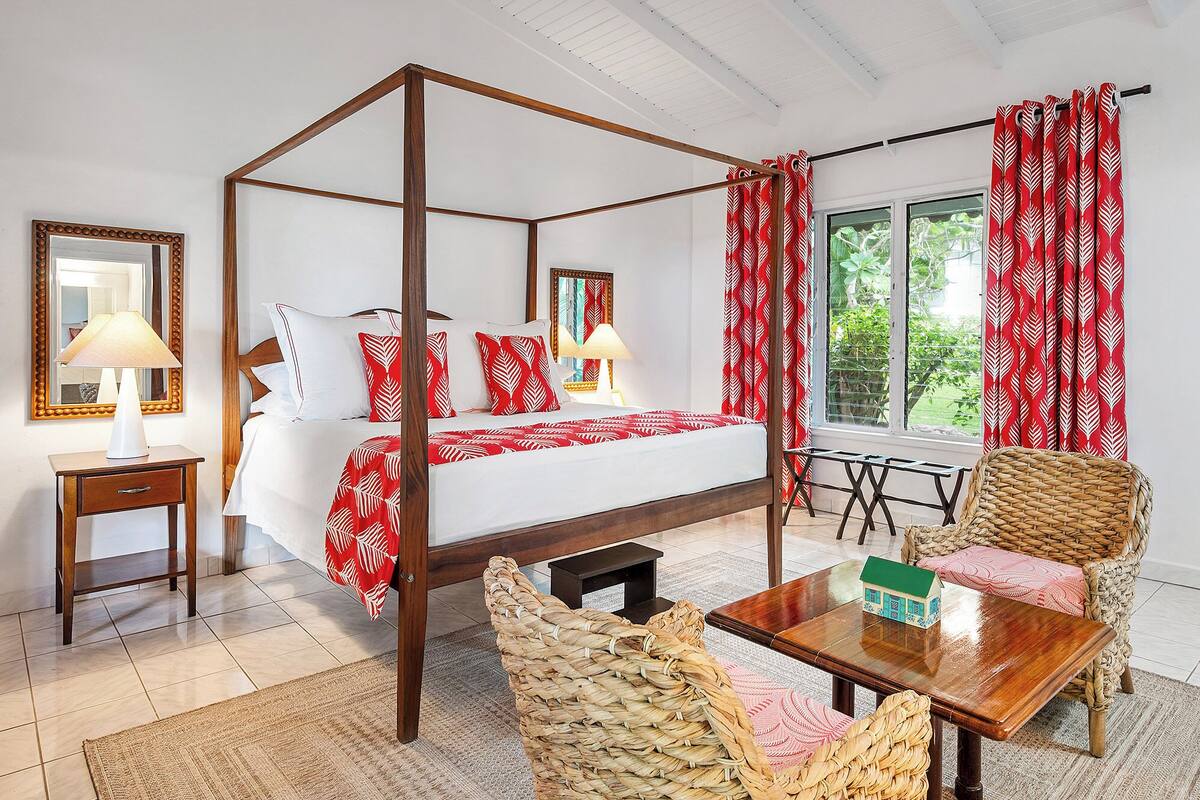 A four-poster bed with a red and white patterned bedspread, a wooden nightstand with a lamp, a wicker chair, a small wooden table, and a view of greenery through the window.