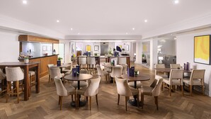 A spacious hotel dining area with multiple round tables and chairs, a central bar area, and a modern kitchen visible in the background.