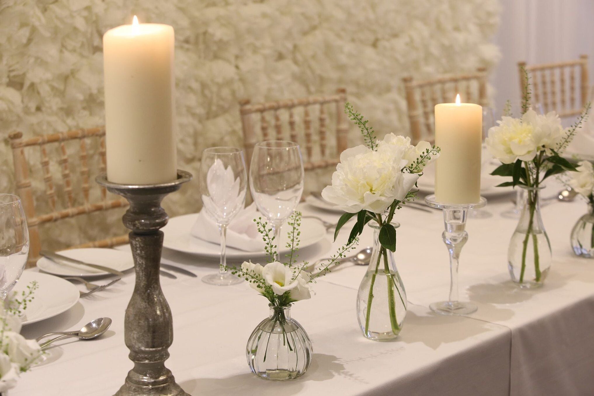 A table set with white plates, clear wine glasses, and a large white candle.
