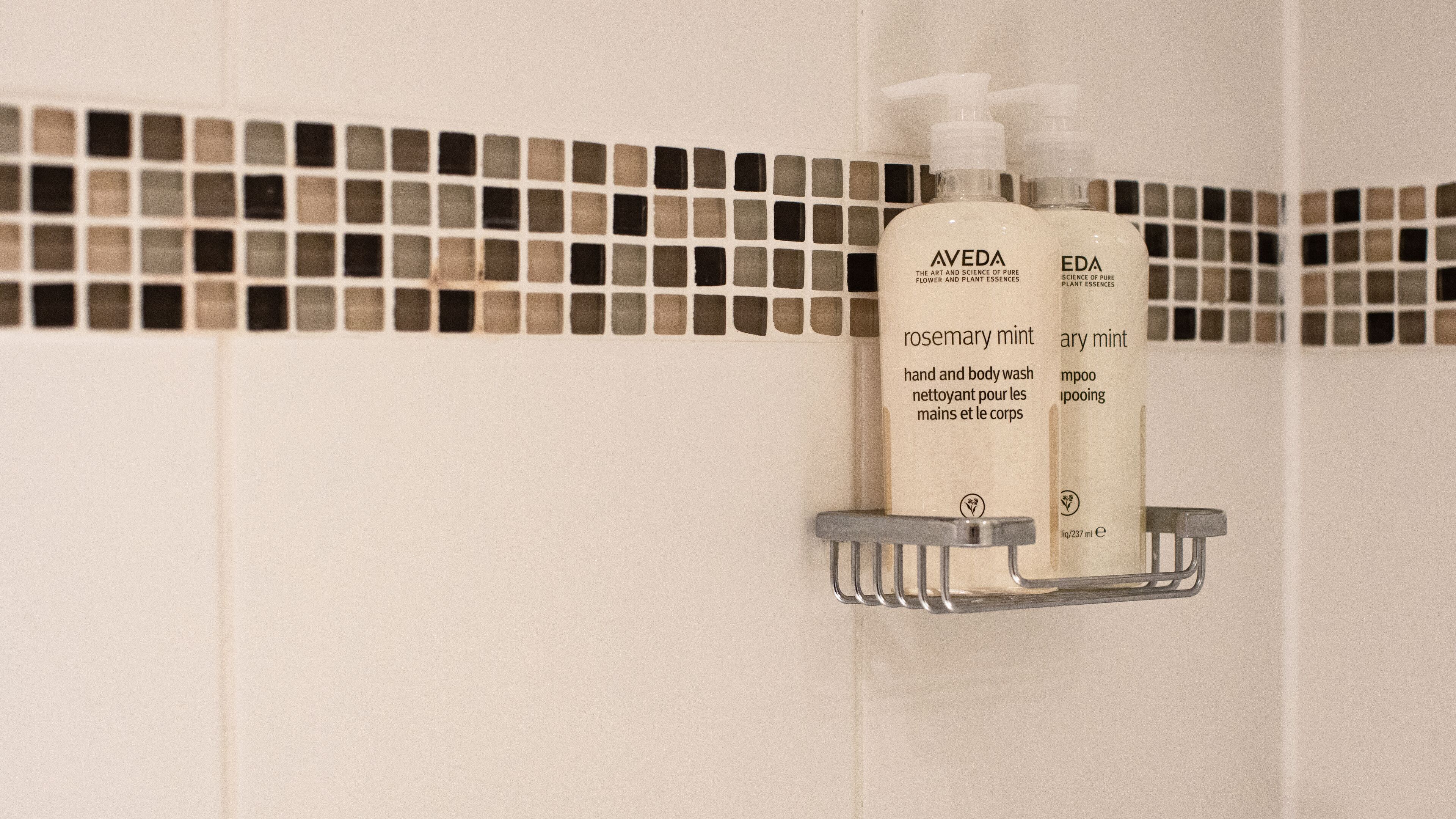 Two bottles of Aveda and EDA hand and body wash on a metal shelf against a tiled wall.