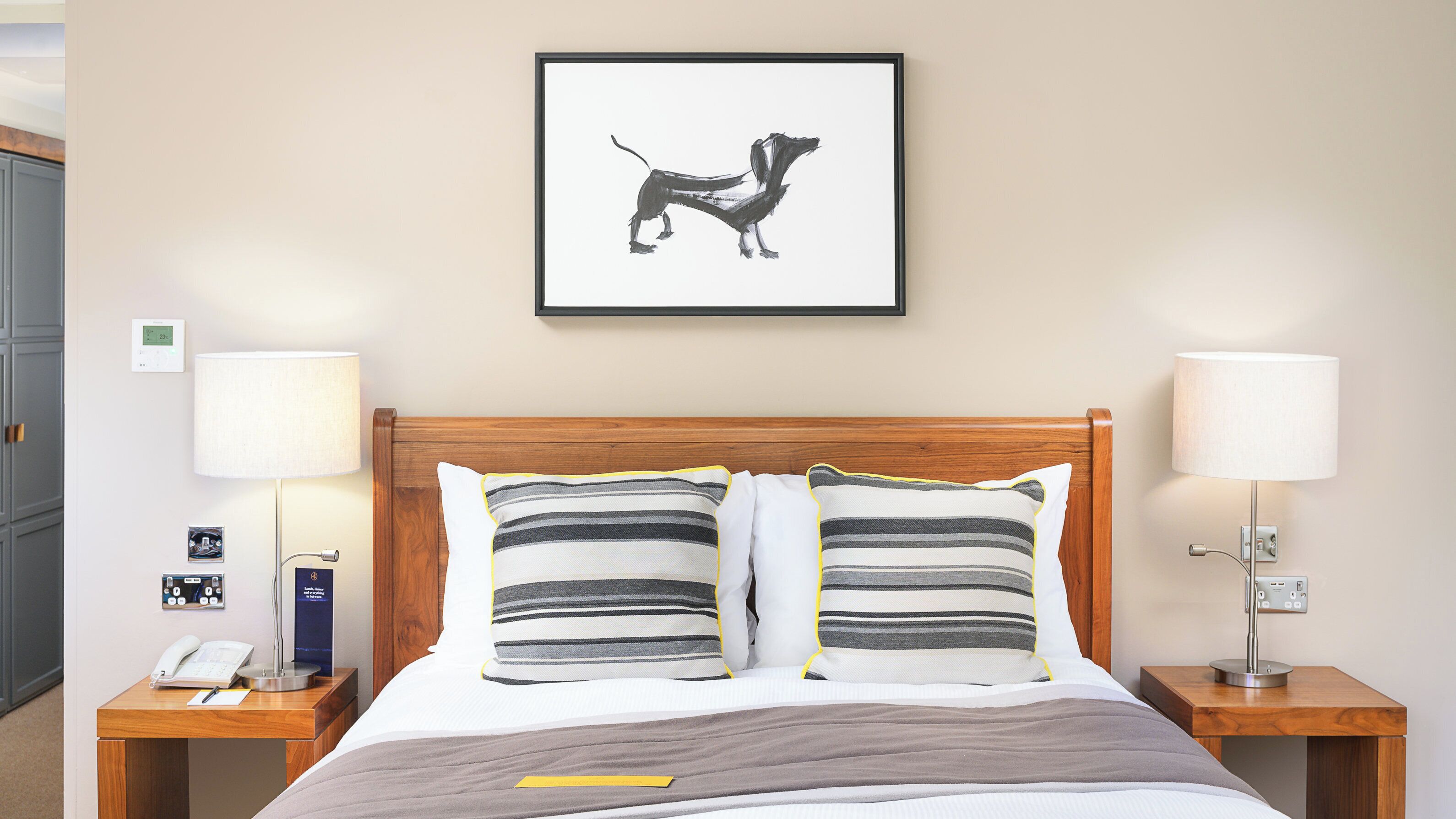 A hotel room with a wooden headboard, two bedside lamps, a framed picture of a dog, a bedside phone, and a bedside table with a lamp and a book.
