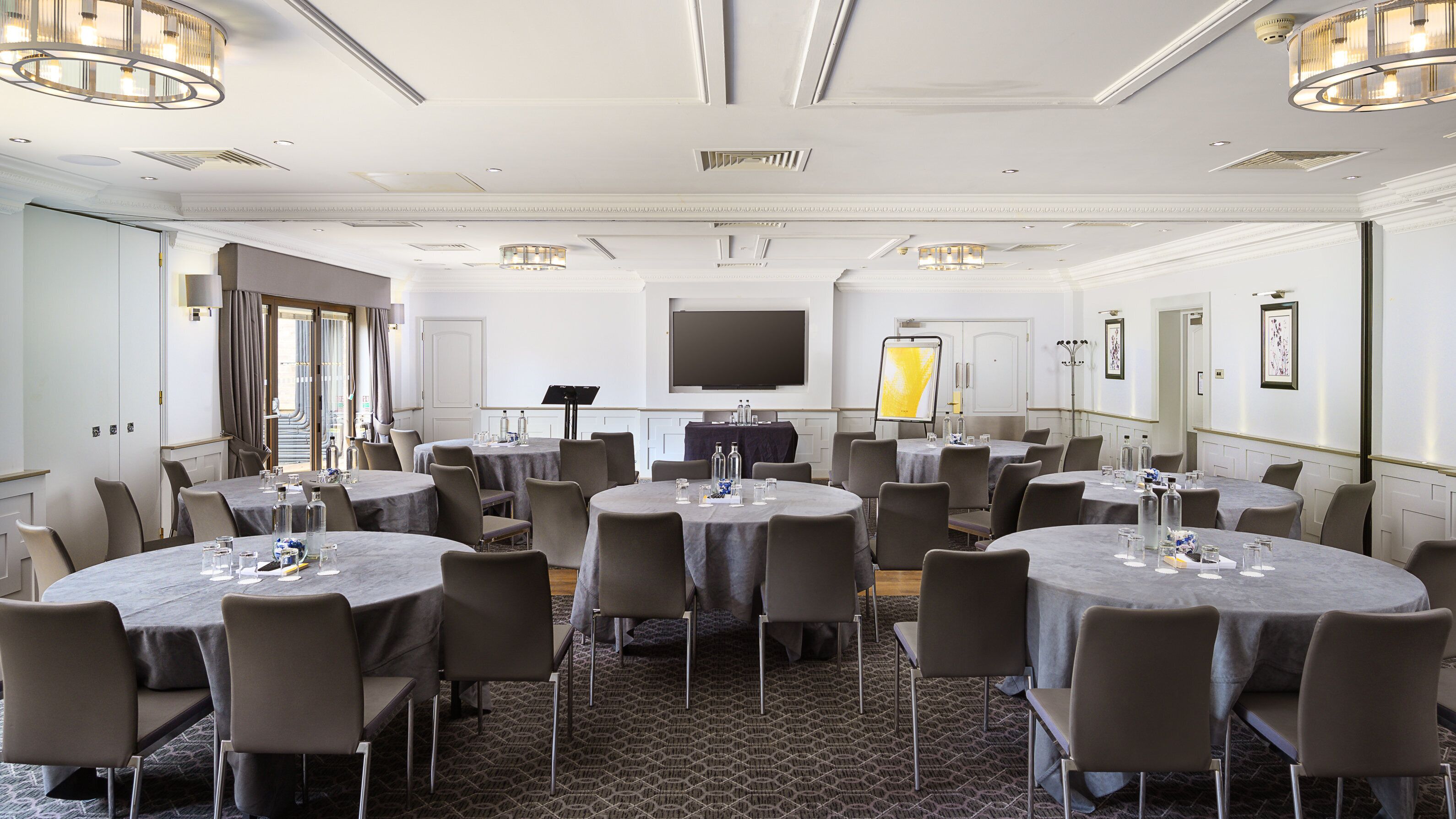 A conference room with multiple round tables, grey chairs, a large flat-screen TV, and a projector screen.