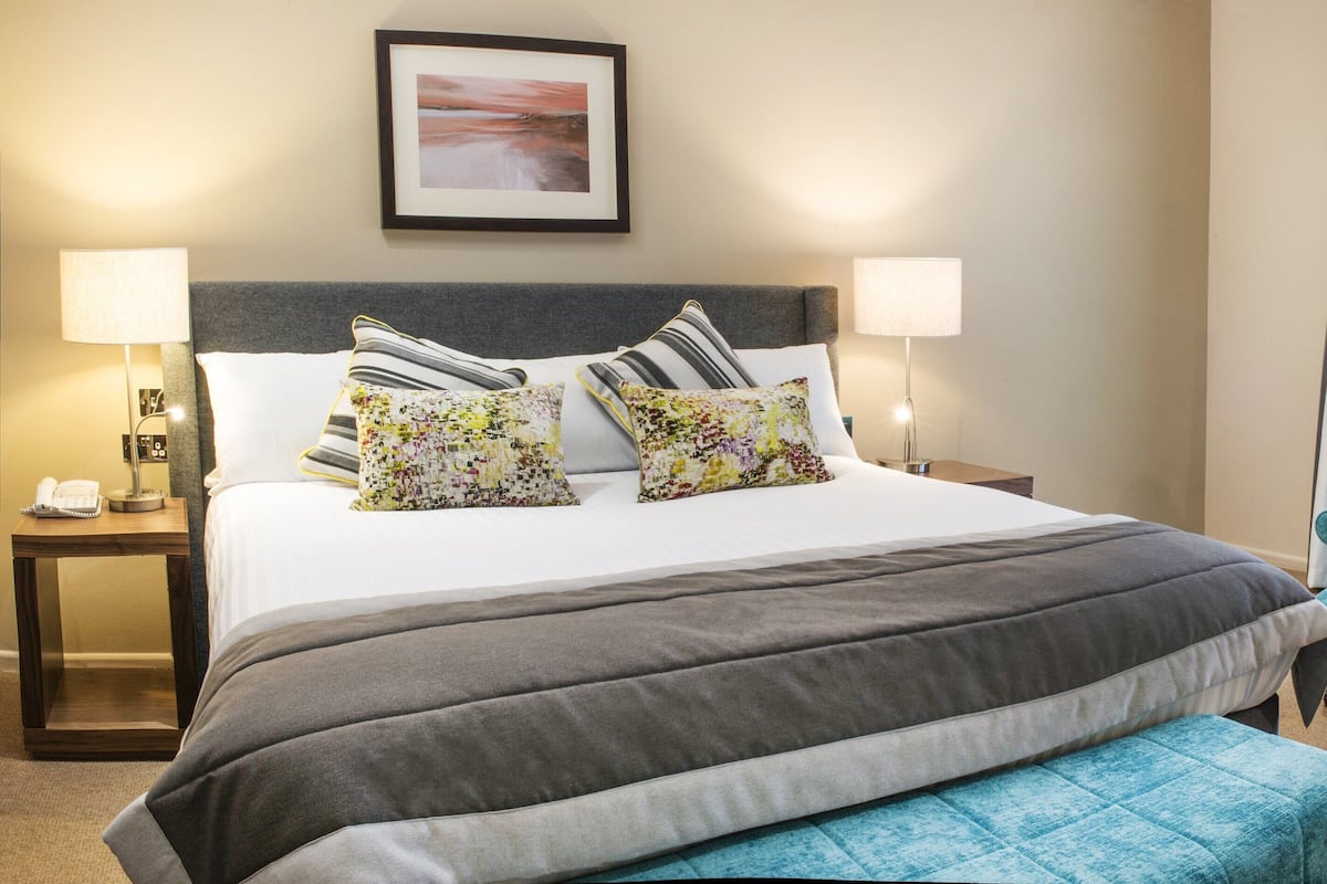A hotel room with a large bed, two bedside lamps, a framed picture on the wall, a blue armchair, and a small round table.