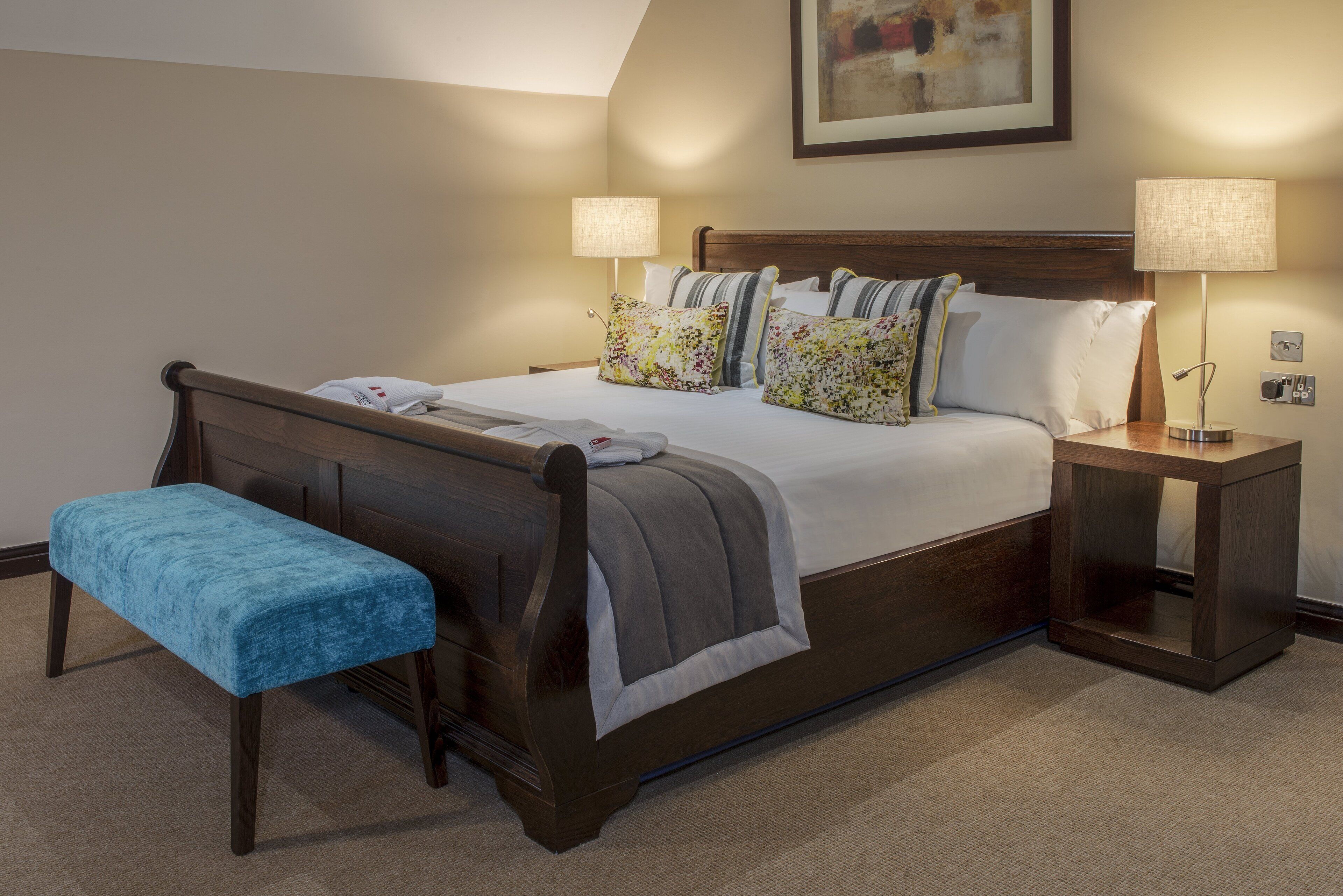 Een slaapkamer met een houten bed, twee nachtkastjes, een bankje en een schilderij aan de muur.