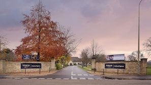 A hotel entrance with a sign for VOCO Oxford Spires, a paved driveway, and a large tree with autumn leaves. - voco Oxford Spires by IHG (Oxford)