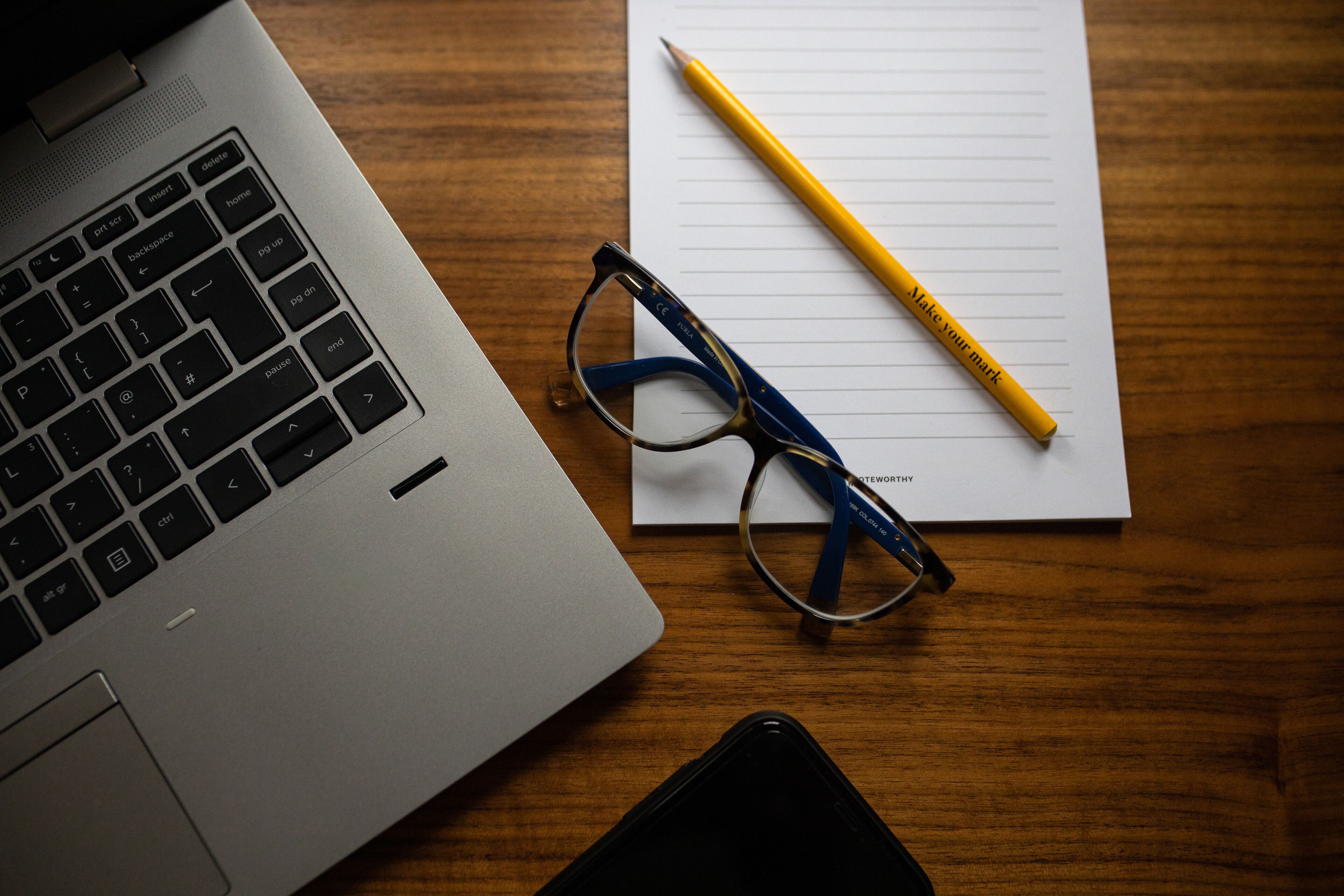 A laptop, a pair of glasses, a pencil, and a smartphone on a wooden table.