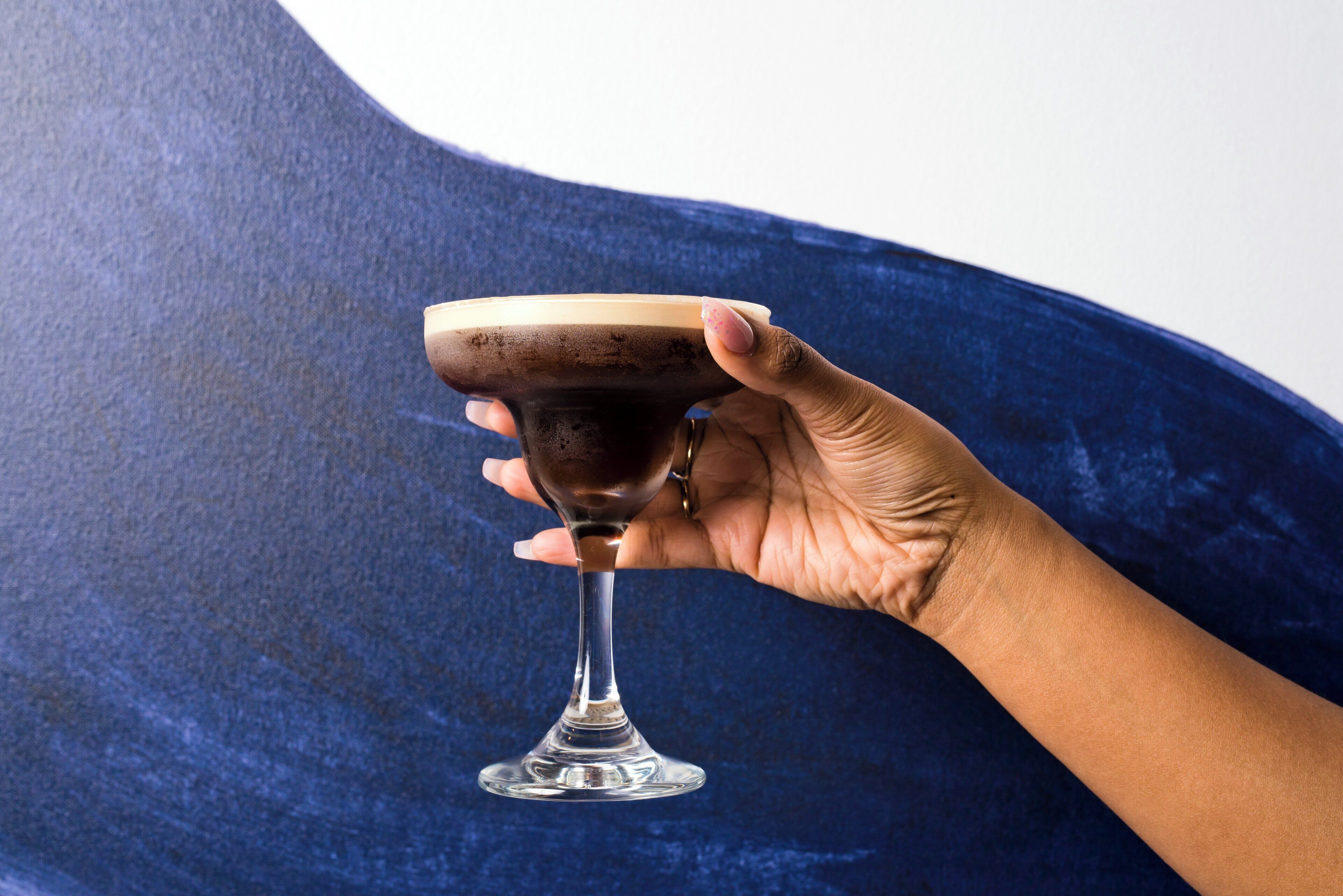 Una mano sosteniendo un vaso con un líquido oscuro, posiblemente un cóctel, sobre un fondo azul con textura.