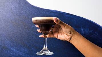 Una mano sosteniendo un vaso con un líquido oscuro, posiblemente un cóctel, sobre un fondo azul con textura.