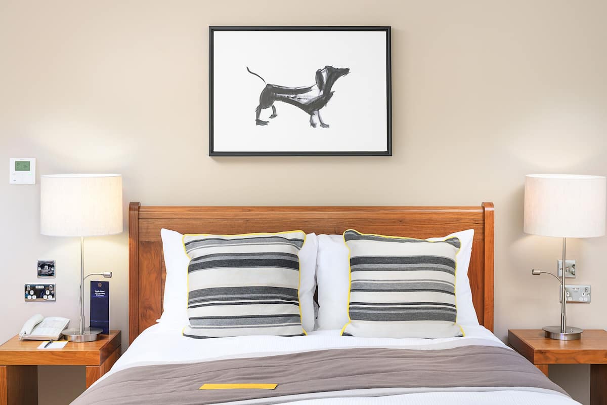 A hotel room with a wooden headboard, two bedside lamps, a framed picture of a dog, a bedside phone, and a digital thermostat.