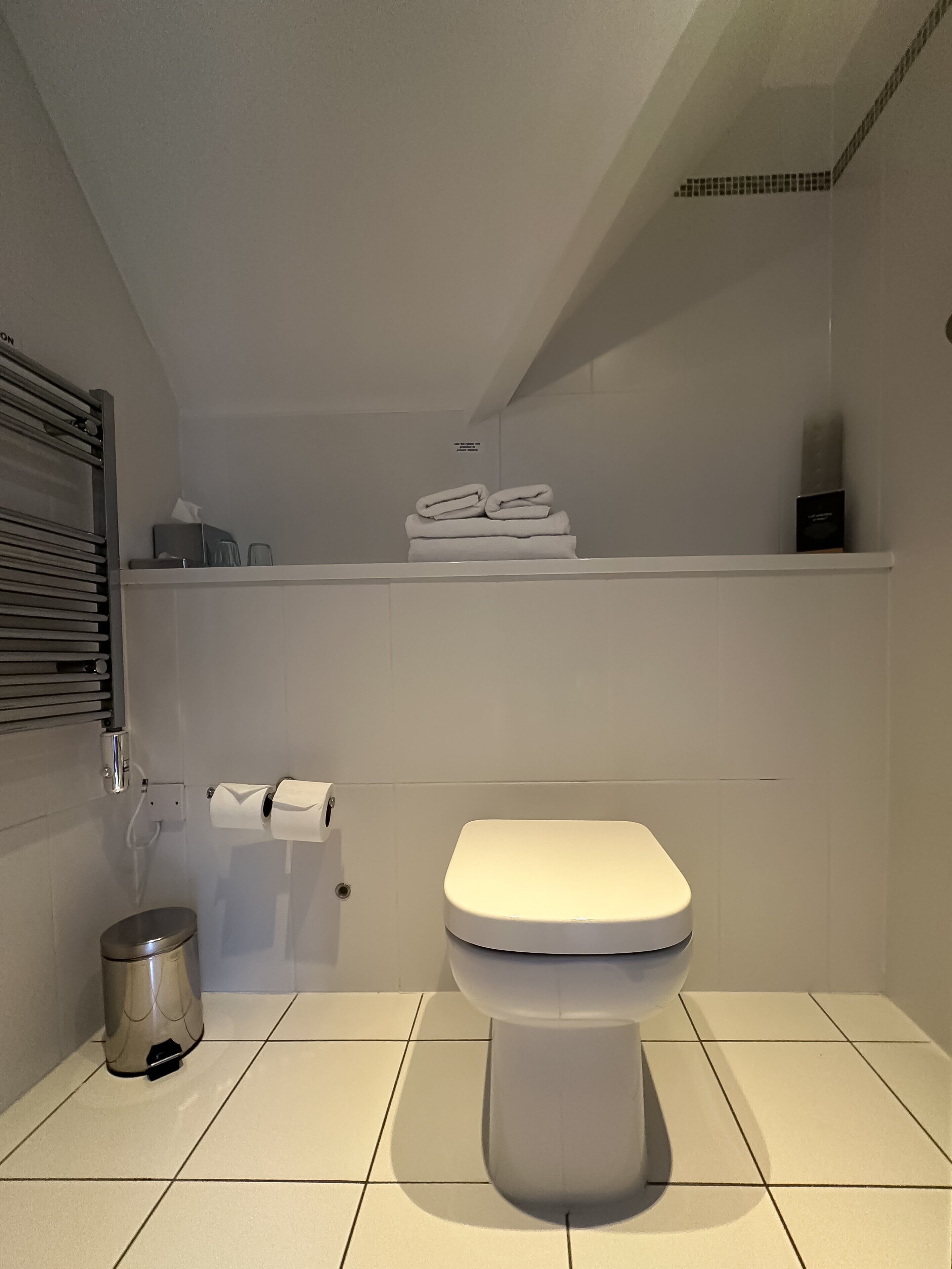 A modern bathroom with a toilet, a trash can, a towel rack, and a shelf with folded towels.