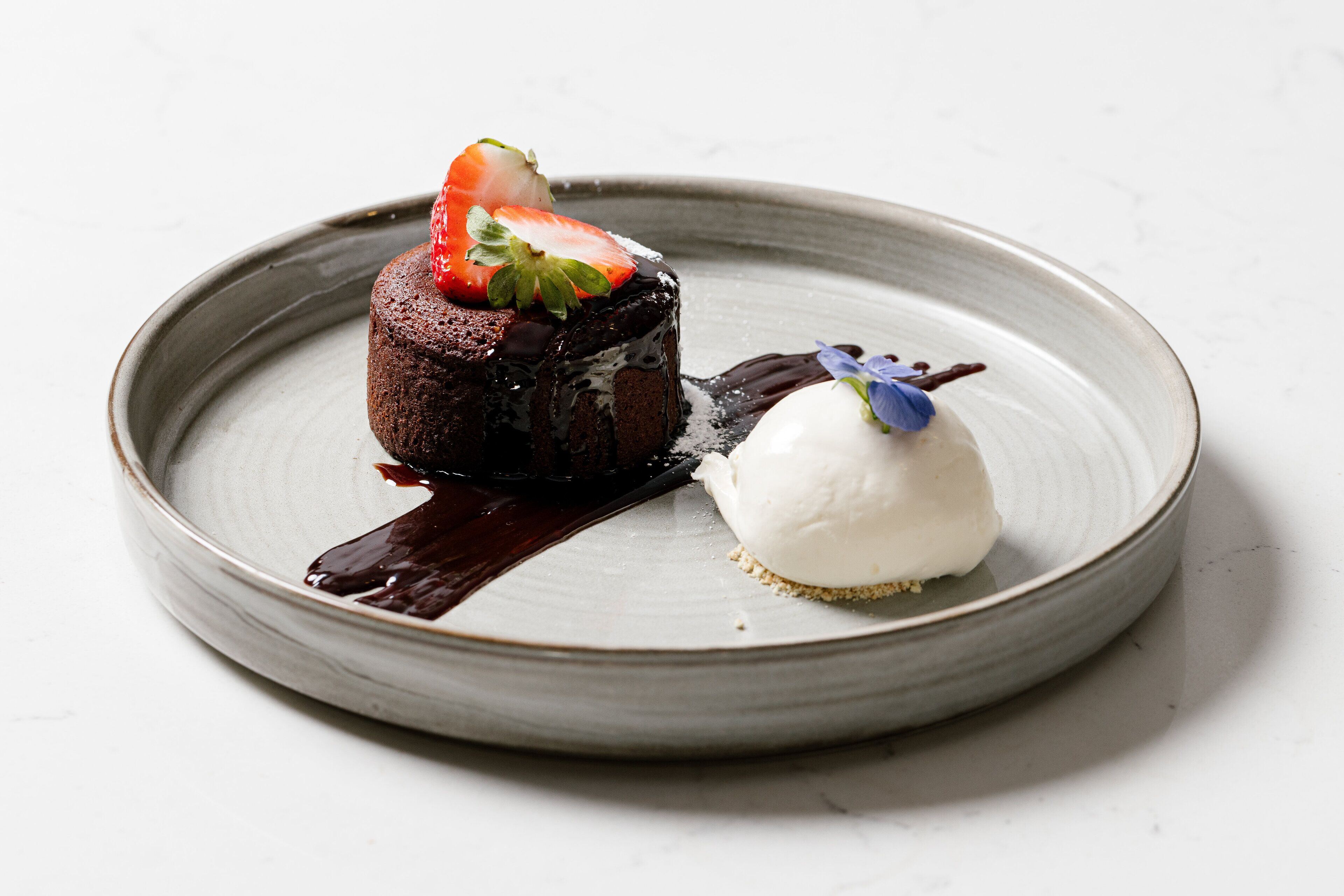A dessert on a ceramic plate with a chocolate cake topped with a strawberry and a scoop of ice cream with a blue flower on top.