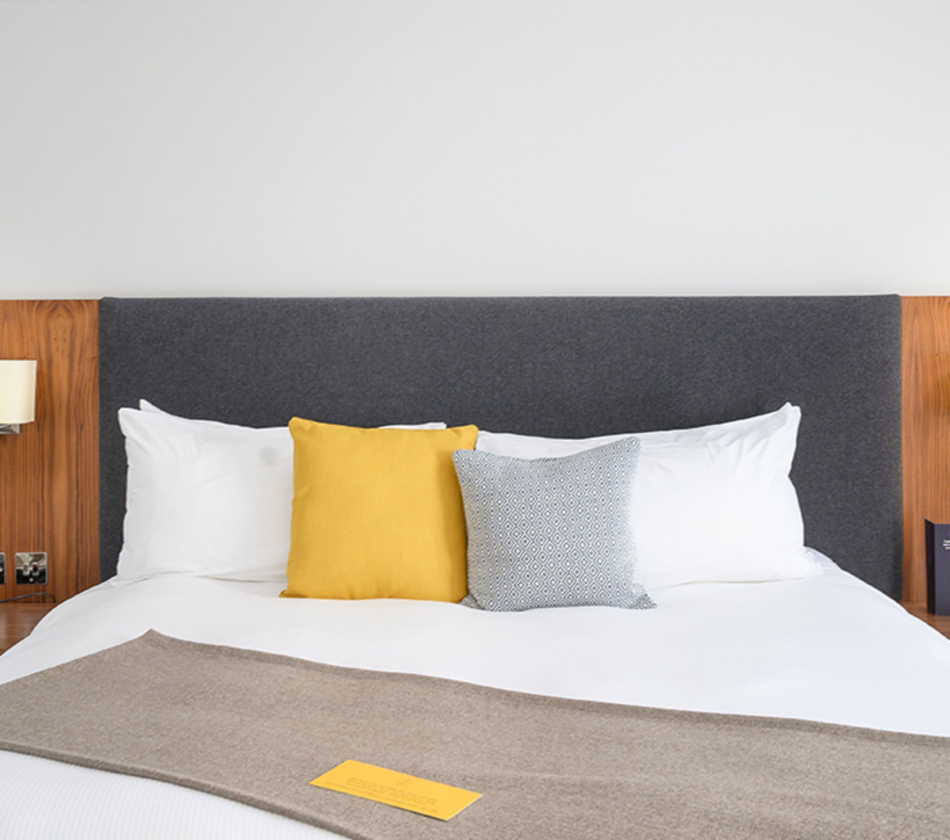 A neatly made bed with white and patterned pillows, a grey headboard, and a brown throw blanket.