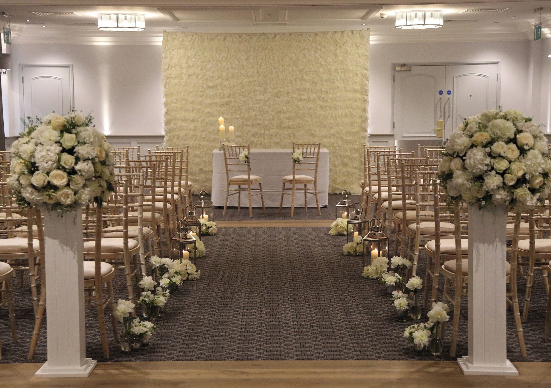 A wedding aisle with white floral arrangements and candles, leading to a table with two chairs.