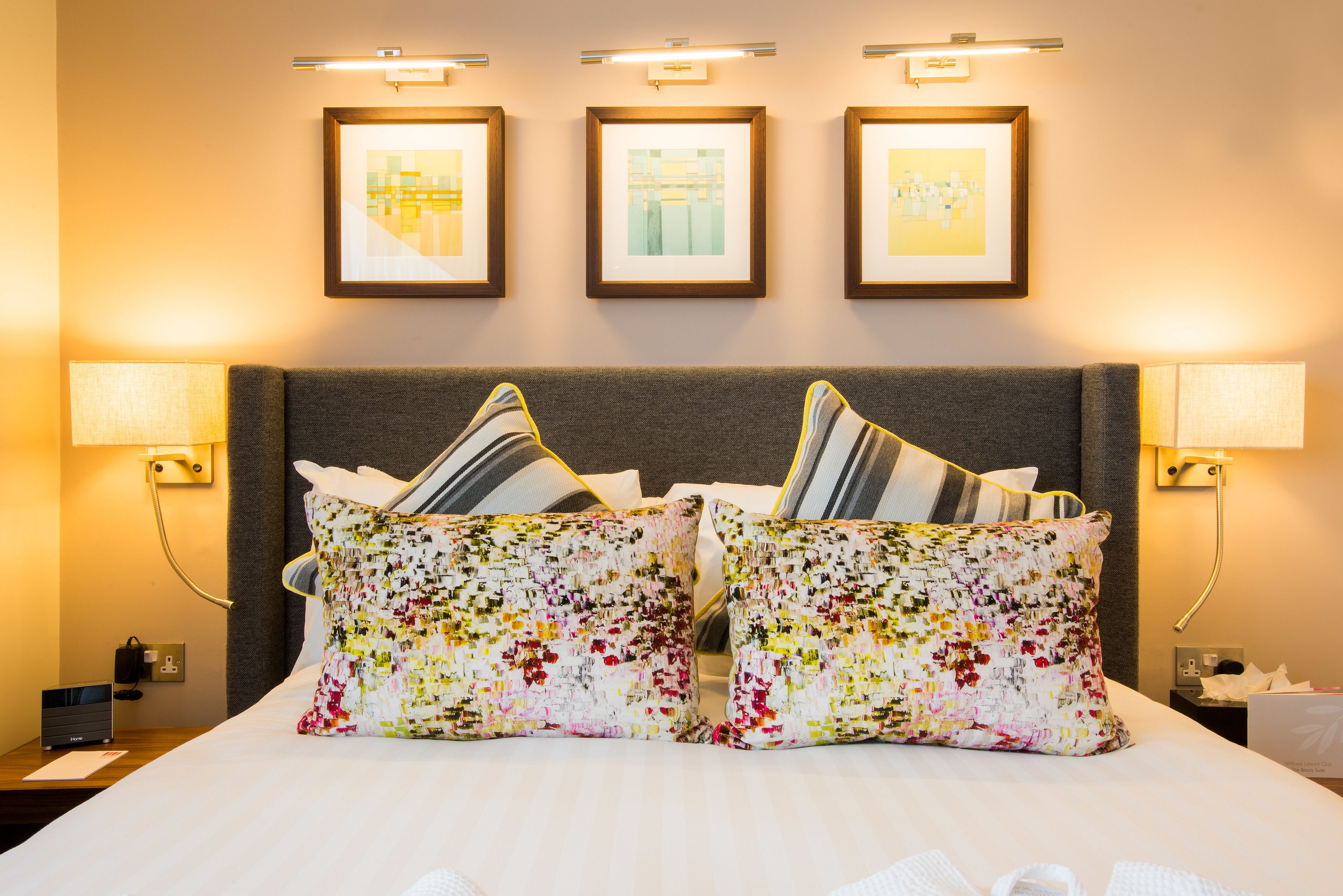 A hotel room with a bed, two pillows, a nightstand, a lamp, and three framed pictures on the wall.