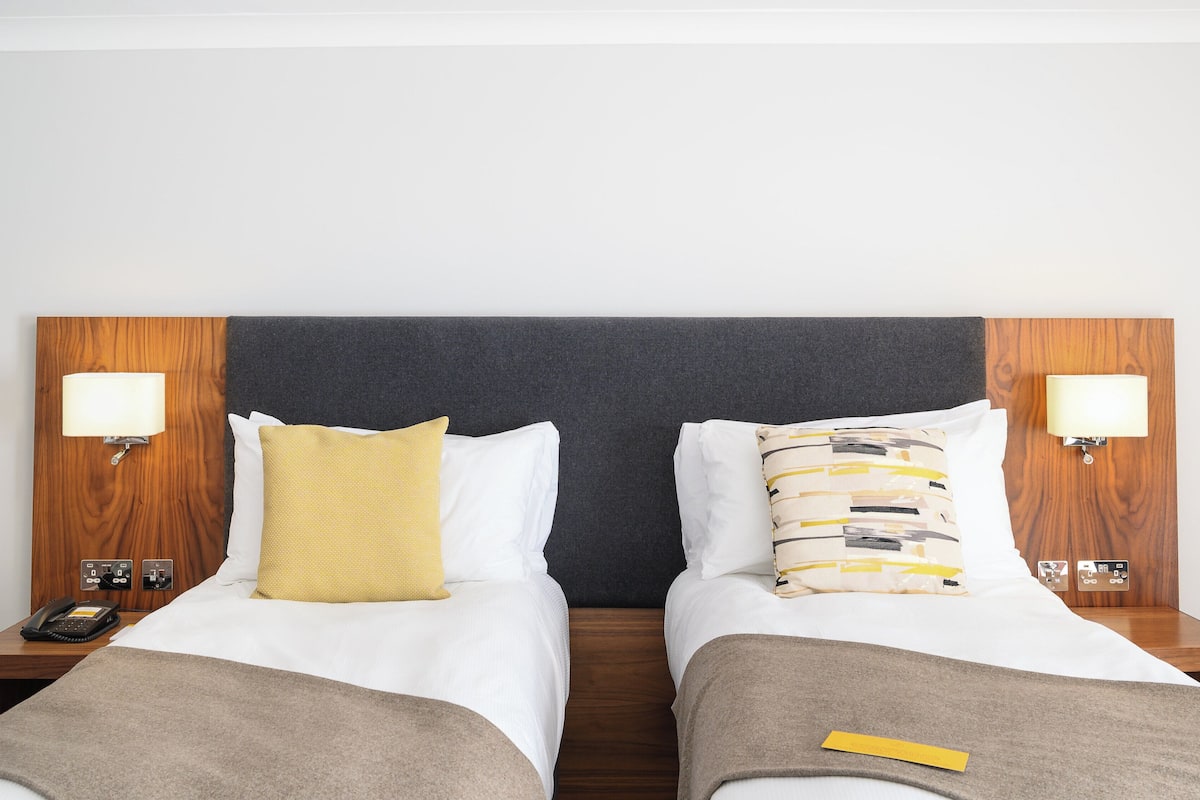 A hotel room with two beds, each with a wooden headboard, white bedding, and a bedside lamp. There is a telephone on the left side of the room.