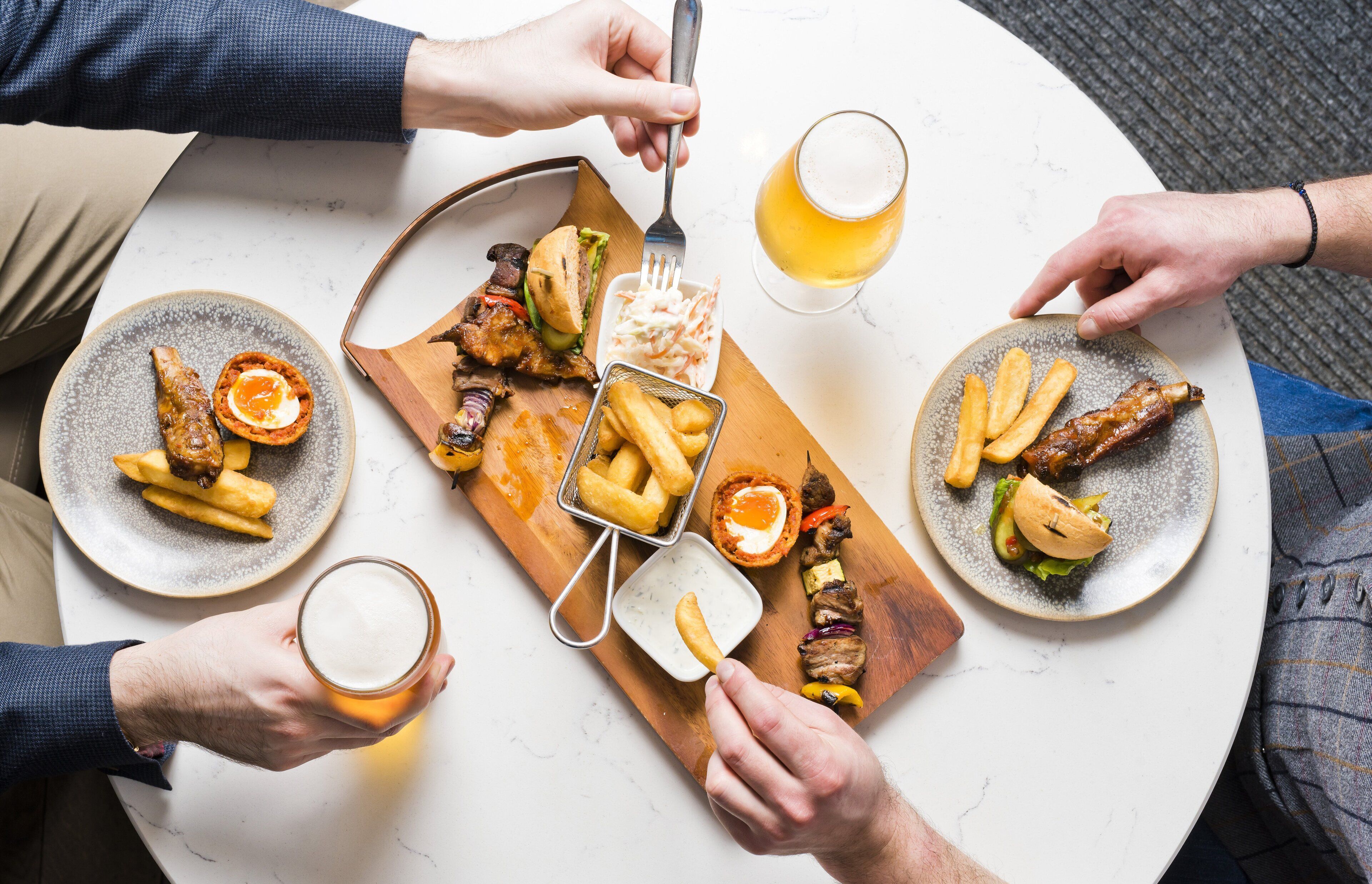 A group of people dining at a round table with various dishes, including meat, vegetables, and fries, and glasses of beer.