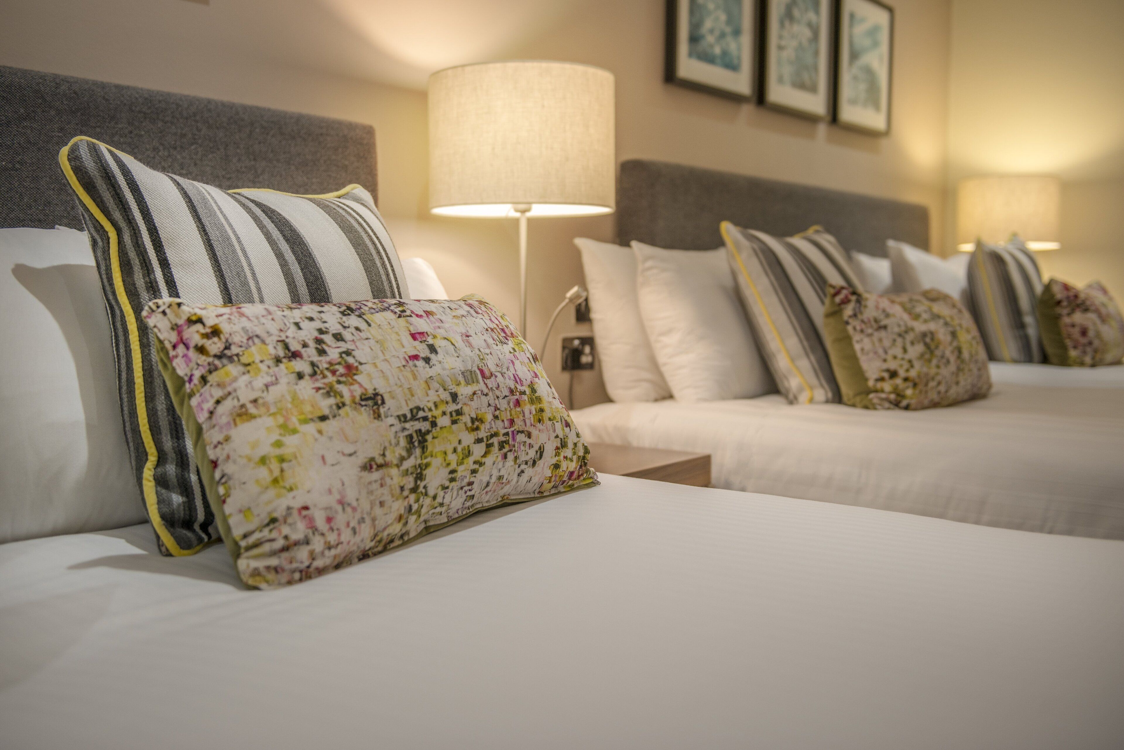 A hotel room with two beds, striped pillows, a floral pillow, and a lamp.