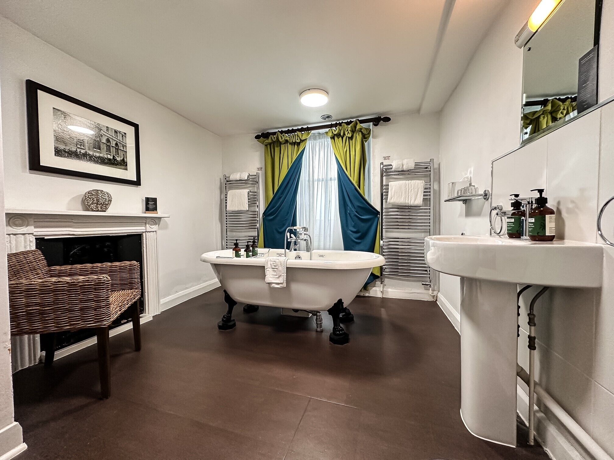 A bathroom with a freestanding bathtub, a sink, a mirror, a wicker chair, and a fireplace.