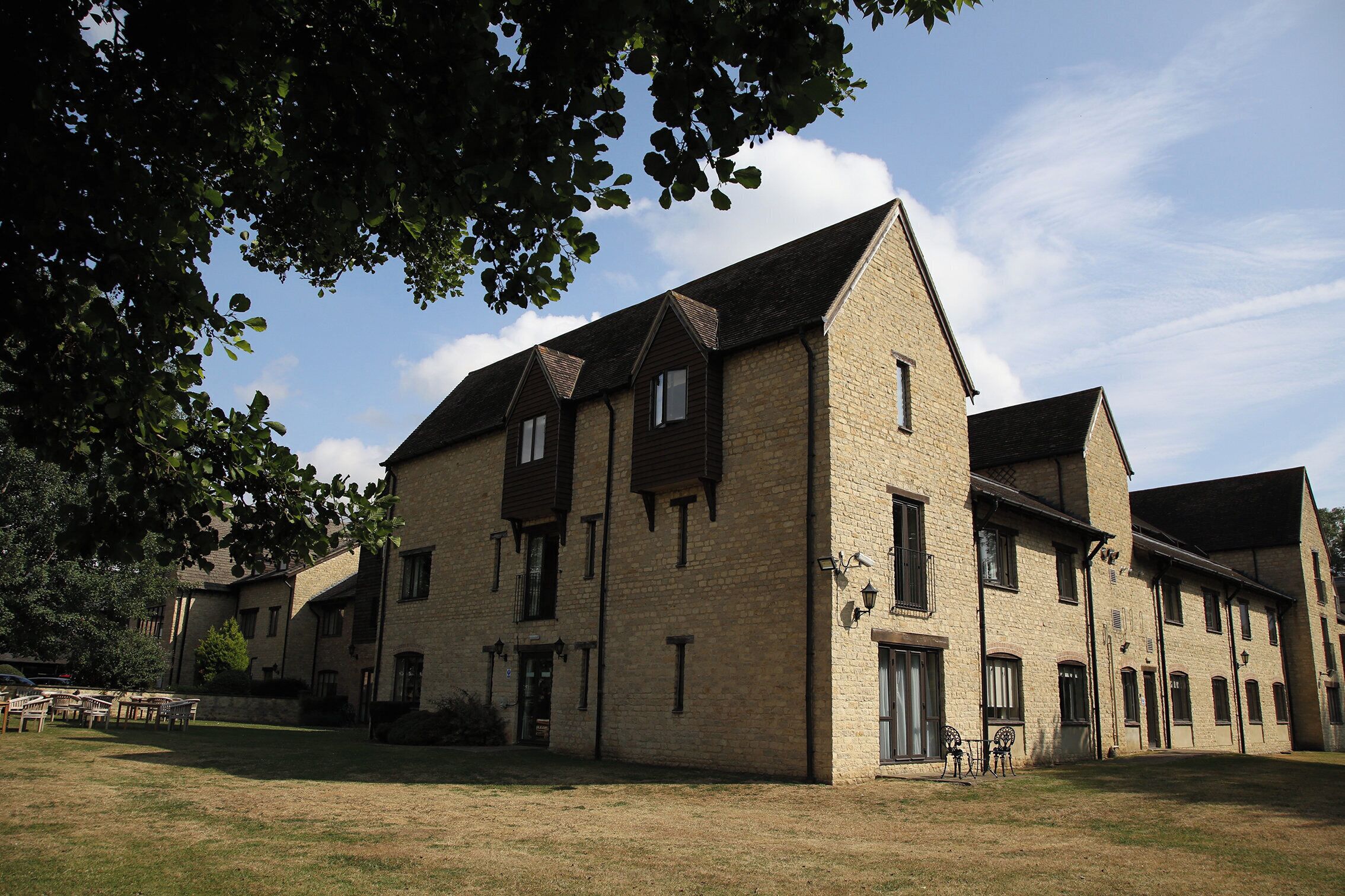 A traditional stone building with multiple windows and a sloped roof, surrounded by a grassy area and trees.