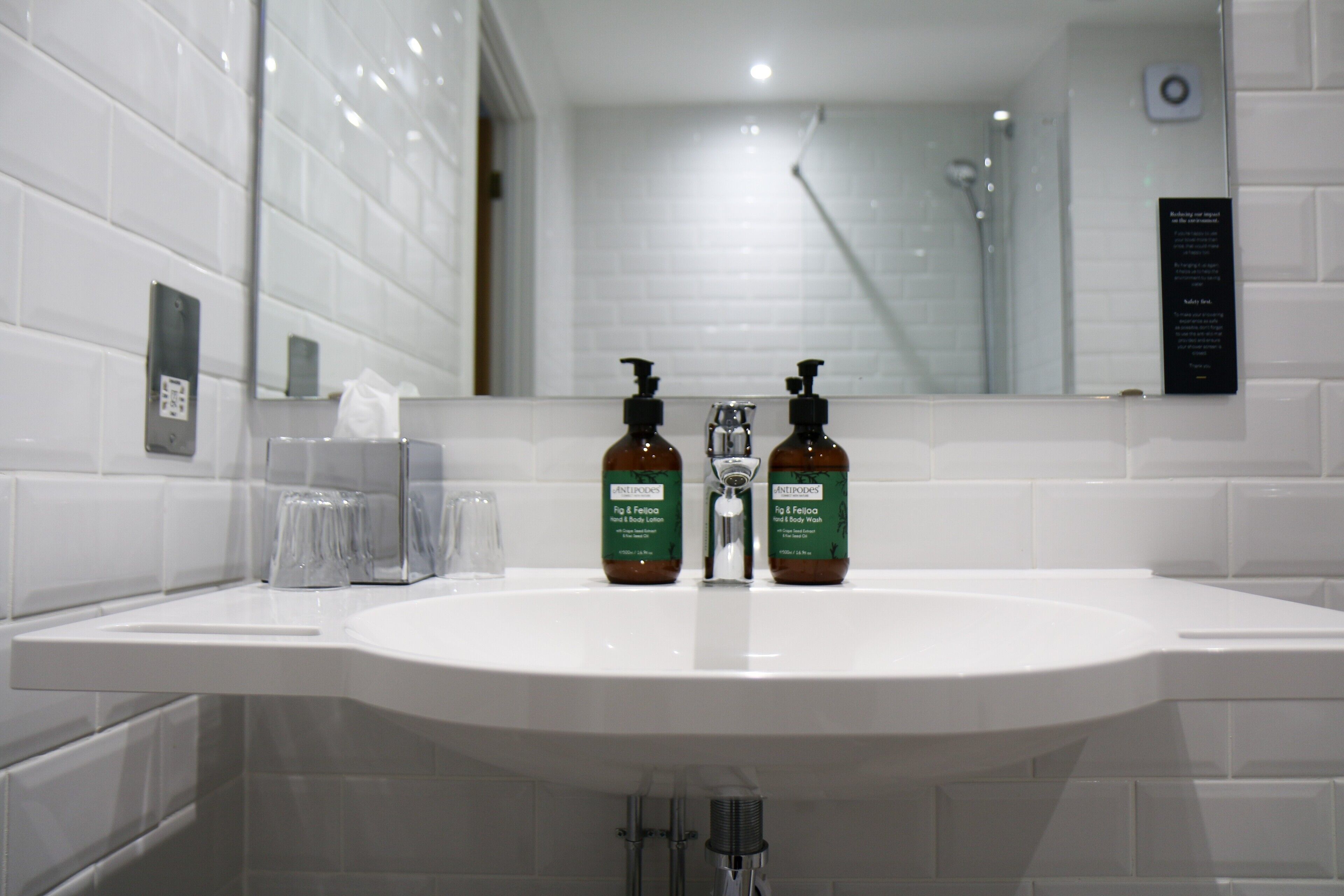 A white bathroom sink with two brown bottles of shampoo and conditioner, a mirror, and a white tiled wall.