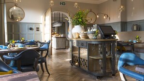 A dining area with round tables, blue upholstered chairs, and a central island with a vase of flowers. - The Painswick (Stroud)