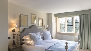 A neatly made bed with white linens and decorative pillows, a bedside table with a lamp, and framed botanical prints on the wall. - The Painswick (Stroud)