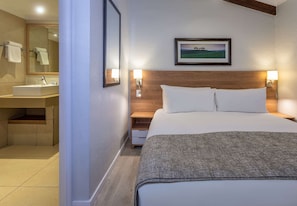 A hotel room with a bed, bedside lamps, a nightstand, a wooden headboard, a framed picture on the wall, and a bathroom with a sink and mirror. - Pine Lake Inn (Mbombela)