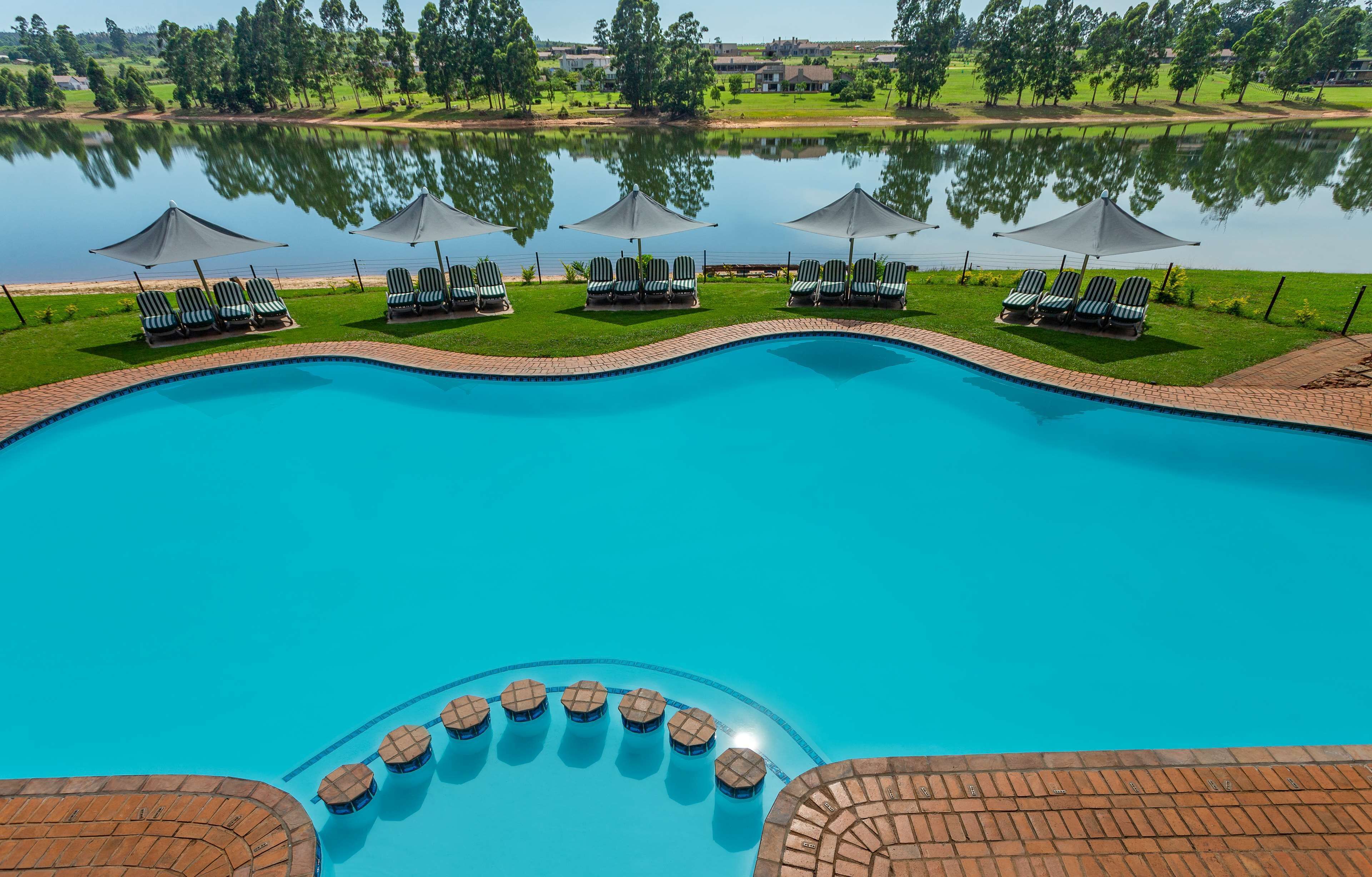 Une piscine entourée d’une terrasse en briques, agrémentée de chaises longues et de parasols, et offrant une vue sur un lac et la verdure environnante.