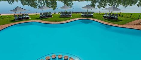 A swimming pool with lounge chairs and umbrellas, surrounded by a brick deck and a view of a lake and greenery.
